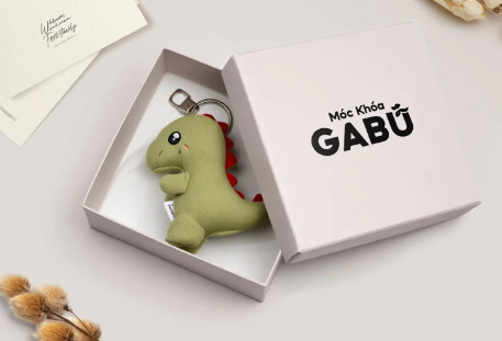 Móc khóa khủng long đặt trong hộp quà nhỏ cùng thiệp tay và hoa khô.