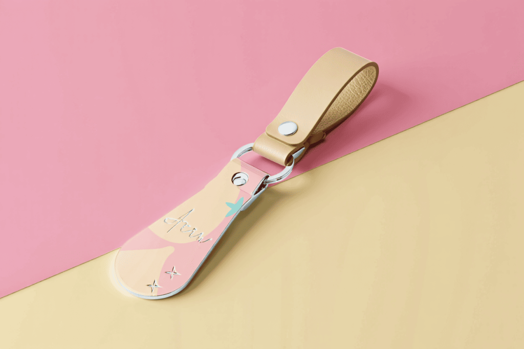 Móc khóa kim loại tối giản viền bo tròn, điểm xuyết màu pastel và dây da be, đặt trên nền hồng kem, ánh sáng dịu, phong cách dễ thương